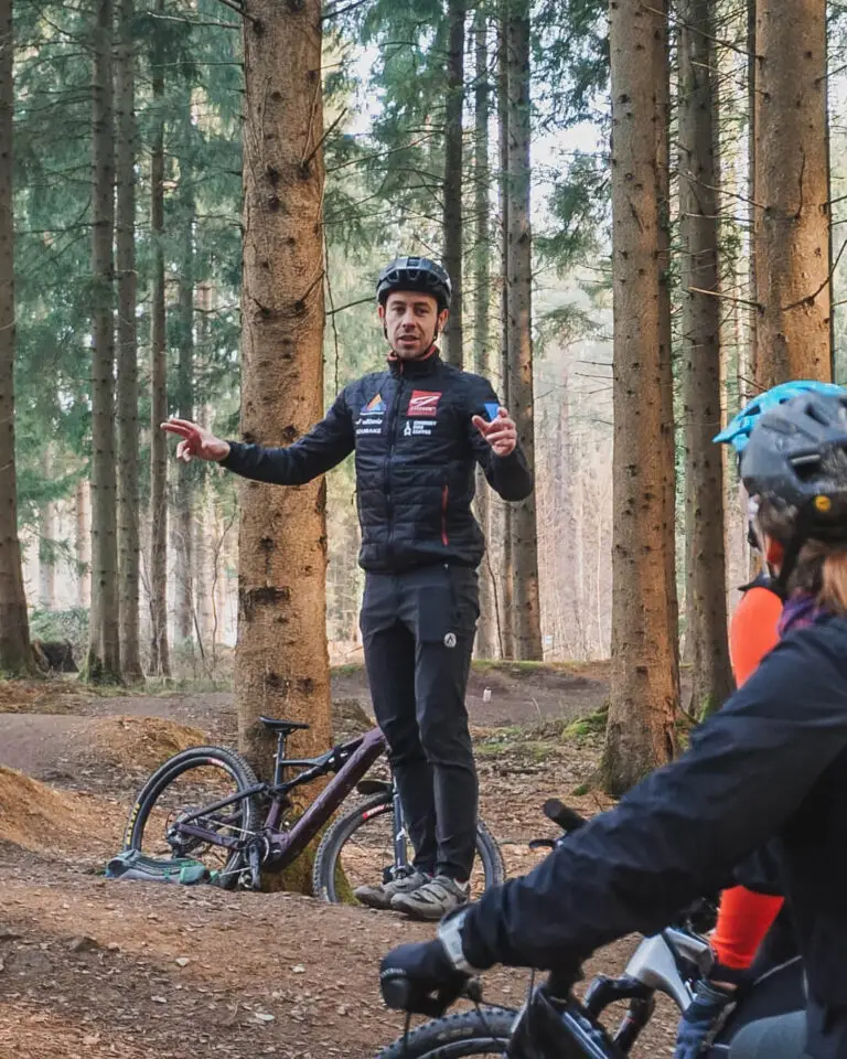 Man in black cycling clothes coaching other MTB riders in a forest, a mountain bike is propped against a tree.