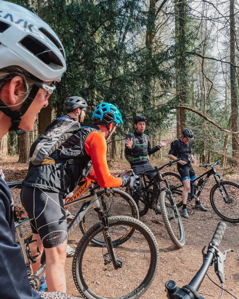 A group of mountain bike riders all listening to a male coach teaching them through a section of track, they are surrounded by trees.